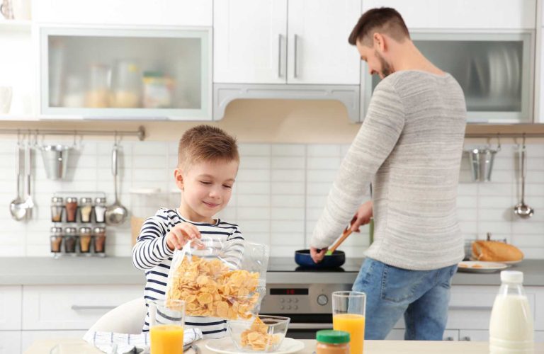 Young boy pouring cereal from a flexible plastic bag into a bowl while his father cooks in the background.