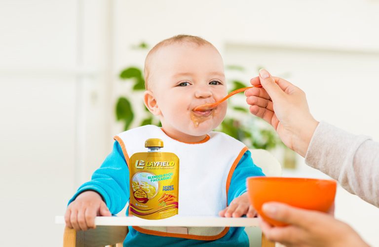 Baby eating from a spoon while sitting in a high chair next to a Layfield-branded spouted pouch of blended baby food.