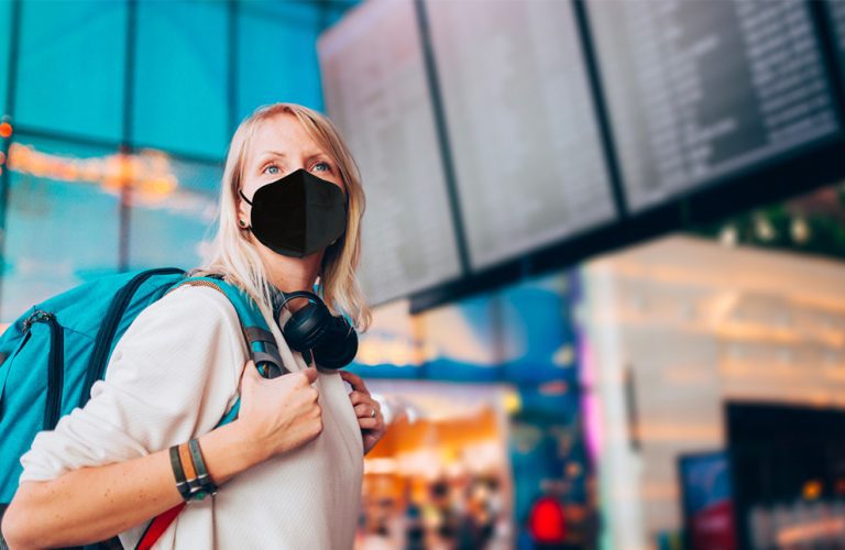 Woman wearing a black protective face mask and backpack while checking a flight board at an airport.
