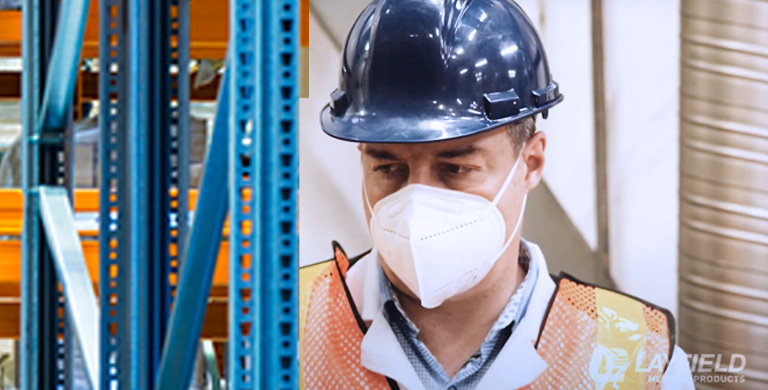 Worker wearing a white respirator mask, safety vest, and hard hat inside a warehouse or industrial facility.