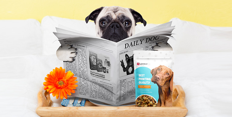 A pug reading a newspaper titled “Daily Dog” beside a Layfield Flexible Films dog food package on a tray with flowers.