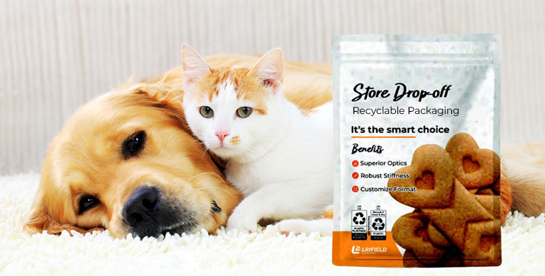 Golden retriever and white cat lying on a carpet next to a pouch of store drop-off recyclable pet treat packaging.