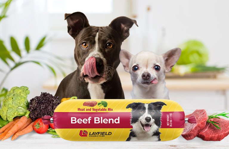 Two dogs behind a packaged raw pet food roll labeled Beef Blend with Layfield branding.