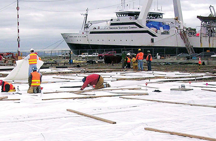 Workers installing vapor barrier sheeting at an industrial construction site near a waterfront.