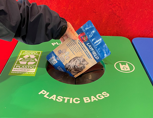 Hand placing a Layfield flexible pouch into a plastic bag Store Drop-Off recycling bin.