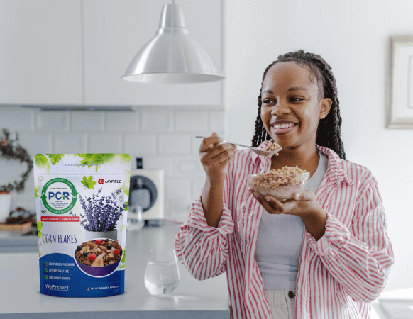 Woman eating cereal in a kitchen with flexible packaging featuring Post-Consumer Recycled (PCR) content displayed in the background.