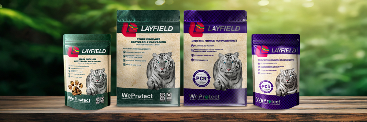 Four Layfield stand-up pouches on a wooden table, featuring recyclable and PCR packaging with tiger graphics, set against a green forest background.